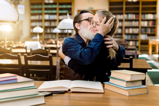 Apuesto hombre barbudo senior abuelo abrazando y besando a su linda nieta, niña con anteojos, sentado a la mesa con muchos libros en la antigua biblioteca