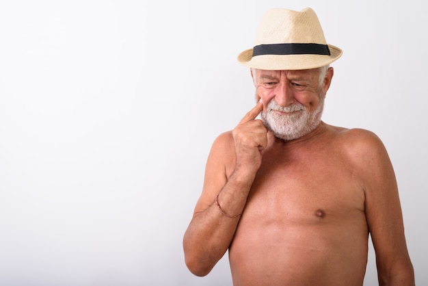 Apuesto hombre barbudo senior pensando mientras mira hacia abajo y con sombrero sin camisa en blanco