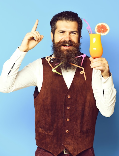 Un apuesto hombre barbudo con tubos coloridos en barba larga y bigote tiene un cabello elegante en la cara sonriente sosteniendo un vaso de cóctel alcohólico en un chaleco de cuero de gamuza vintage en el fondo azul del estudio