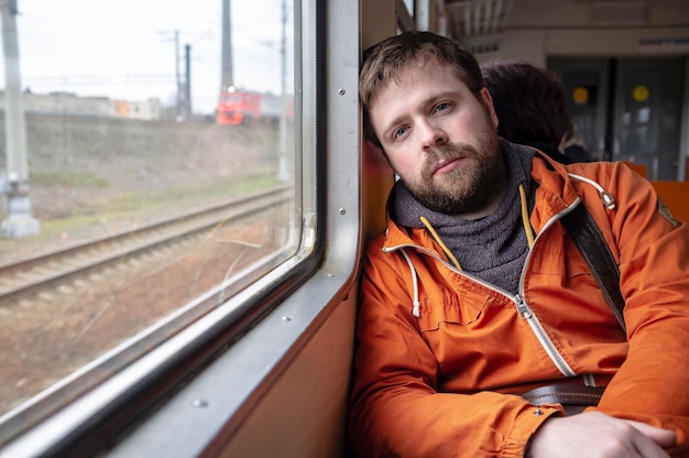 Un apuesto hombre barbudo viaja en el tren apoyado contra la pared cerca de la ventana por fatiga