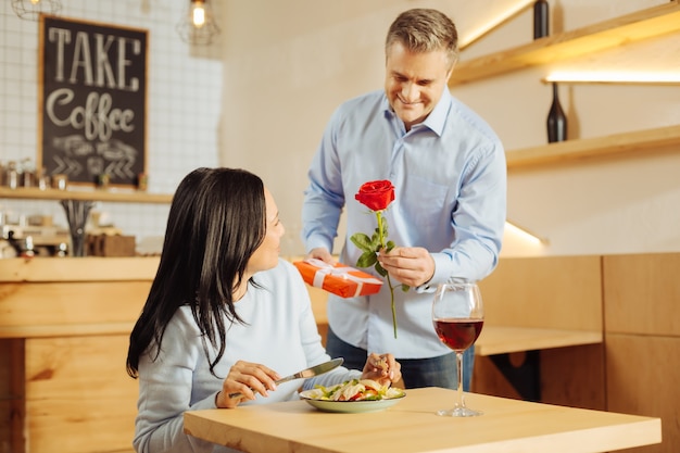 Apuesto hombre bien construido inspirado sonriendo y dando una rosa y un regalo a su bonita mujer morena contenta mientras ella tiene una cena romántica