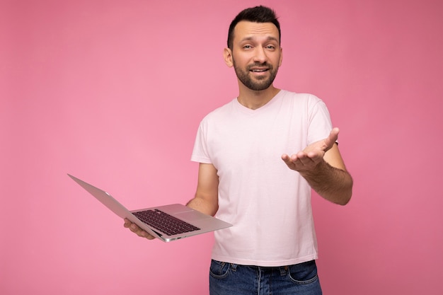 Apuesto hombre brunet preguntando sosteniendo la computadora portátil y mostrando la mano mirando a la cámara en camiseta en