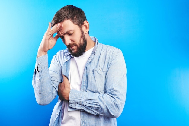 Un apuesto hombre caucásico sostiene la cabeza, experimenta estrés constante, dolor de cabeza y agotamiento emocional en el trabajo, trabajador de oficina aislado en un fondo azul con espacio para copiar