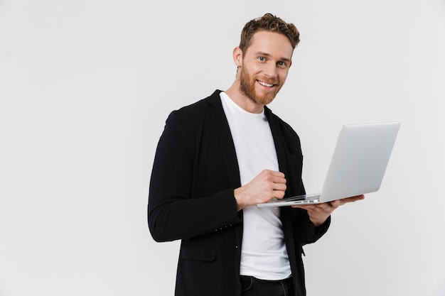 Apuesto hombre complacido en chaqueta sonriendo y usando laptop aislado sobre pared blanca