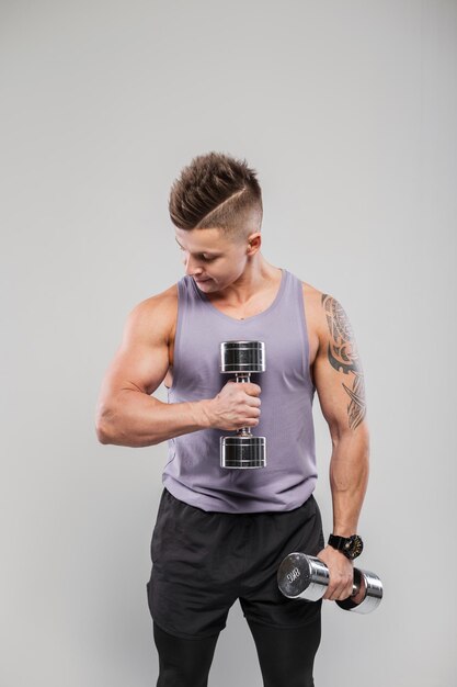 Apuesto hombre deportivo fuerte con peinado en entrenamiento de ropa deportiva con pesas sobre un fondo gris Deportes y estilo de vida saludable