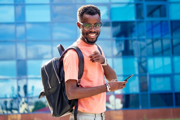 Apuesto hombre empollón afroamericano de moda con gafas en ropa elegante camiseta colorida con una mochila en los hombros de pie contra el fondo de las ventanas azules de la universidad