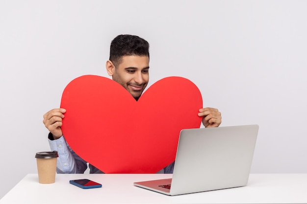 Un apuesto hombre feliz sentado en el escritorio sosteniendo un corazón rojo y hablando por videollamada en una laptop sonriendo expresando sentimientos románticos teniendo citas en línea en un estudio interior aislado en fondo blanco