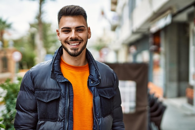 Apuesto hombre hispano con barba sonriendo feliz y confiado en la ciudad con abrigo de invierno