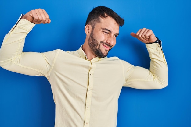 Apuesto hombre hispano de pie sobre fondo azul mostrando los músculos de los brazos sonriendo orgulloso concepto de fitness