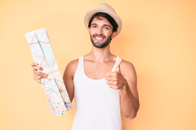 Apuesto hombre hispano sosteniendo un mapa de la ciudad para un viaje de verano sonriendo feliz y positivo pulgar hacia arriba haciendo excelente y señal de aprobación