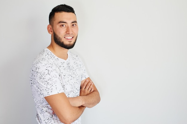 Apuesto hombre kazajo dentudo sonríe, mira a la cámara con las manos cruzadas sobre fondo blanco. Retrato de joven feliz asiático barbudo vestido casual aislado en estudio