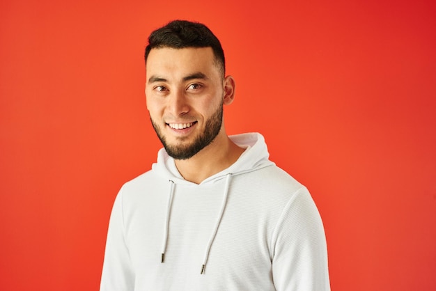 Apuesto hombre kazajo con dientes sonrisas sobre fondo rojo. Retrato de joven feliz asiático barbudo vestido de blanco casual aislado en estudio