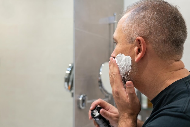 Apuesto hombre maduro aplicando espuma de afeitar en el rostro para cortarse la barba y el bigote con navaja