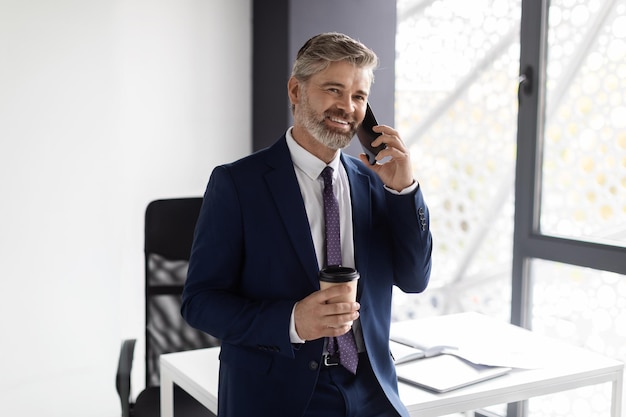 Apuesto hombre de mediana edad hablando por teléfono celular y bebiendo café en la oficina