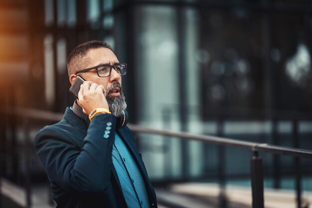 Foto un apuesto hombre de mediana edad va a trabajar y habla por teléfono inteligente frente al distrito de oficinas.