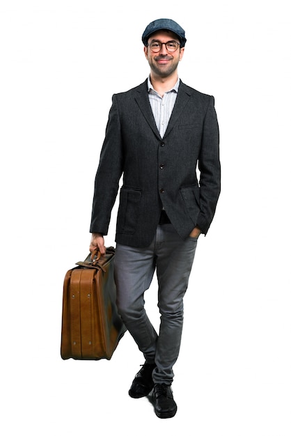 Apuesto hombre moderno con boina y gafas caminando y sosteniendo un maletín vintage