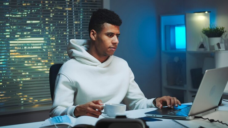 Foto apuesto hombre multirracial bebiendo una taza de café y trabajando en la computadora por la noche hay rascacielos ...