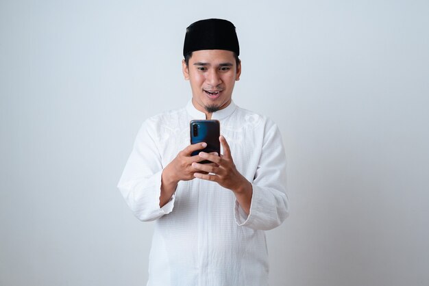 Apuesto hombre musulmán vestido con ropa musulmana y sosteniendo su teléfono charlando contra la pared blanca
