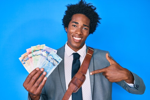 Apuesto hombre de negocios afroamericano con cabello afro sosteniendo dólares canadienses apuntándose con el dedo a uno mismo sonriendo feliz y orgulloso