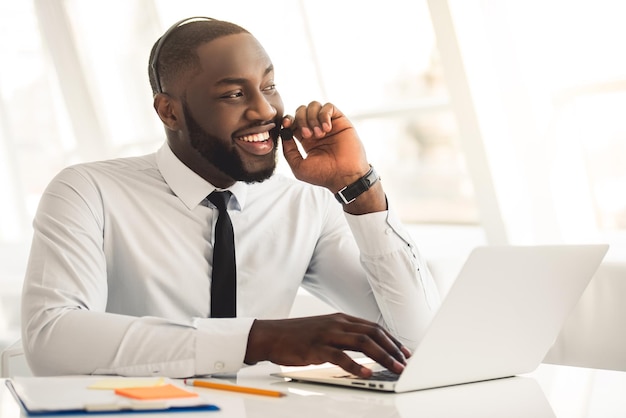 Un apuesto hombre de negocios afroamericano con traje y auriculares habla y sonríe mientras trabaja con una laptop en la oficina