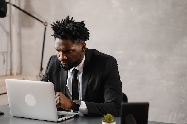 Apuesto hombre de negocios afroamericano en traje clásico usa laptop y sonríe mientras trabaja en la oficina