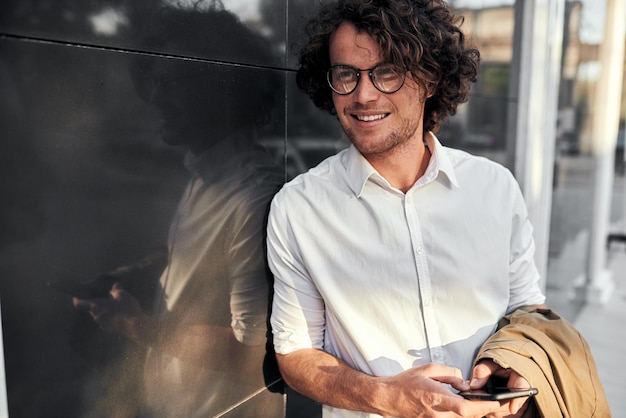 Foto apuesto hombre de negocios apoyado en una pared mientras está de pie al aire libre y usa un teléfono inteligente en la calle hombre feliz con cabello rizado con camisa blanca y abrigo beige en la mano mirando a sus amigos