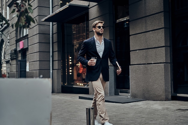 Apuesto hombre de negocios. Apuesto joven en ropa casual elegante con vaso desechable mientras camina por la calle de la ciudad