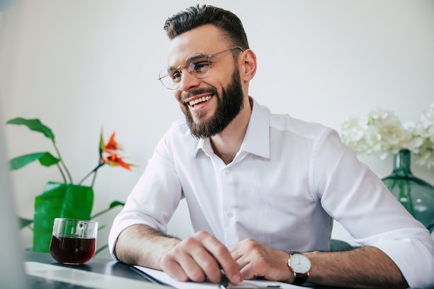 Apuesto hombre de negocios barbudo con gafas y una camisa blanca está trabajando en la computadora portátil y tiene una conversación web en línea con su equipo.