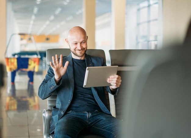 Apuesto hombre de negocios calvo barbudo sonriente en traje comunicarse con alguien de la tableta en el salón del aeropuerto