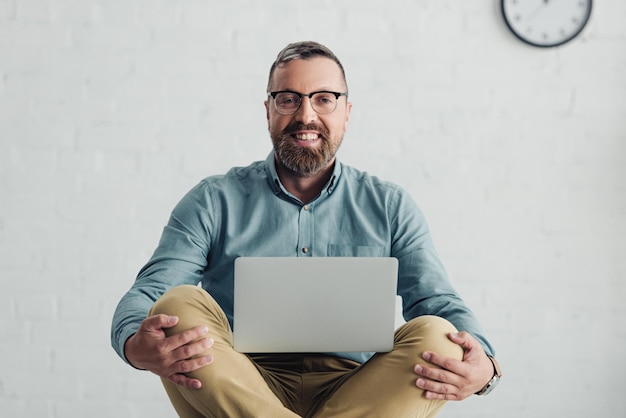 Apuesto hombre de negocios en camisa y gafas sonriendo y sosteniendo la computadora portátil