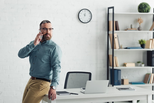 Apuesto hombre de negocios en camisa hablando por teléfono inteligente y mirando hacia otro lado