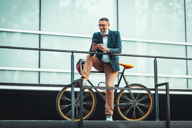 Un apuesto hombre de negocios casual de mediana edad va a la oficina en bicicleta. Está sentado en la bicicleta y usando un teléfono inteligente.