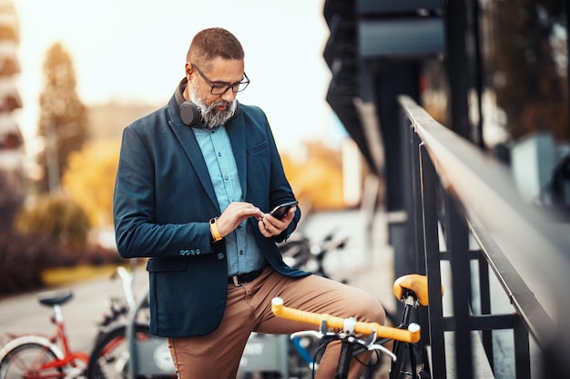 Un apuesto hombre de negocios casual de mediana edad va a trabajar en bicicleta. Está parado en la bicicleta y usando un teléfono inteligente.