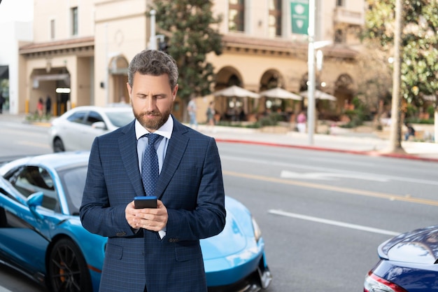 Apuesto hombre de negocios charlando en el teléfono inteligente stand by auto de lujo al aire libre. coche disponible. hombre en traje caro cerca del automóvil. negocio de seguros de automóviles. distribuidor exitoso. administrador seguro de alquiler en línea