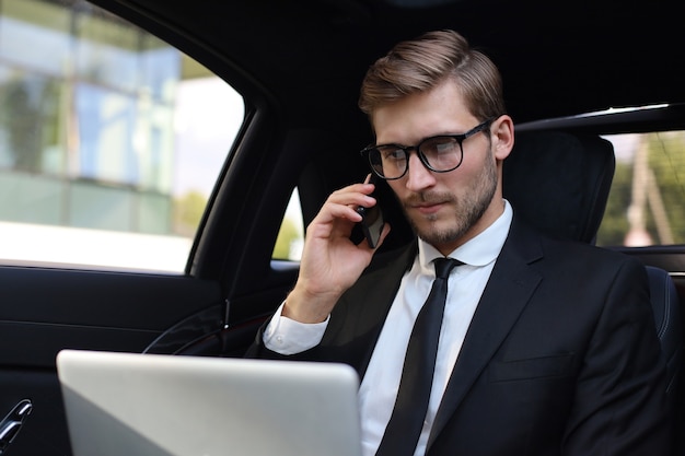 Apuesto hombre de negocios confiado en traje hablando por teléfono inteligente y trabajando con ordenador portátil mientras está sentado en el coche.