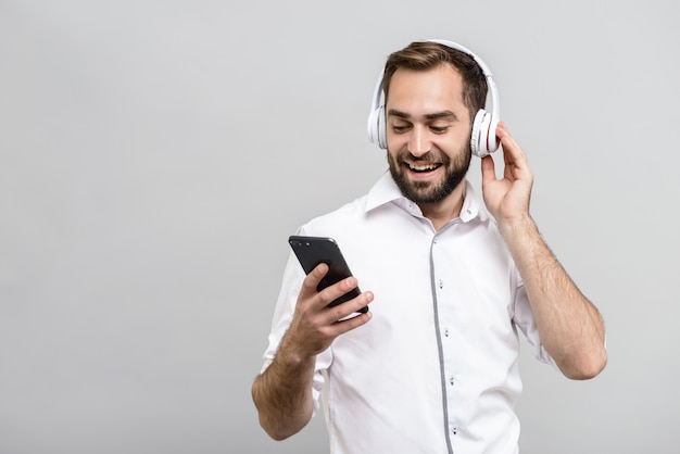 Apuesto hombre de negocios confiado vistiendo traje que se encuentran aisladas sobre la pared gris, escuchando música con auriculares y teléfono móvil