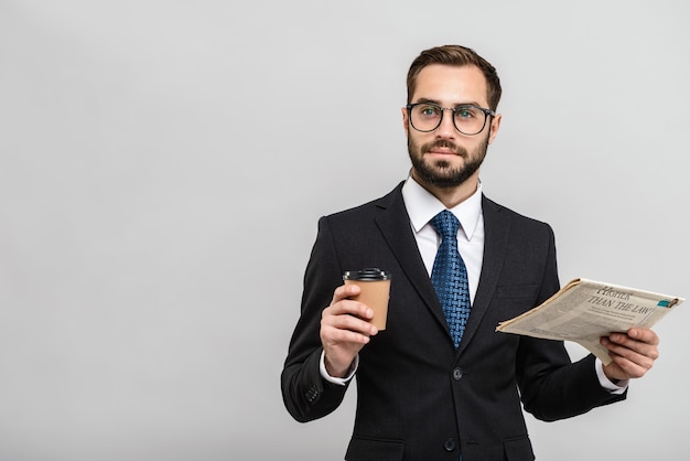 Apuesto hombre de negocios confiado vistiendo traje que se encuentran aisladas sobre la pared gris, leyendo el periódico, bebiendo café para llevar