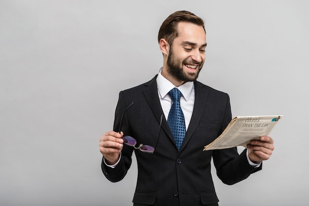 Apuesto hombre de negocios confiado vistiendo traje que se encuentran aisladas sobre la pared gris, leyendo el periódico, usando anteojos