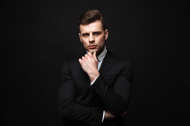 Foto apuesto hombre de negocios confiado vistiendo traje que se encuentran aisladas sobre la pared negra, posando