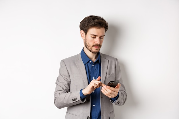 Apuesto hombre de negocios en elegante traje charlando en el teléfono inteligente, mediante teléfono móvil, de pie sobre fondo blanco.