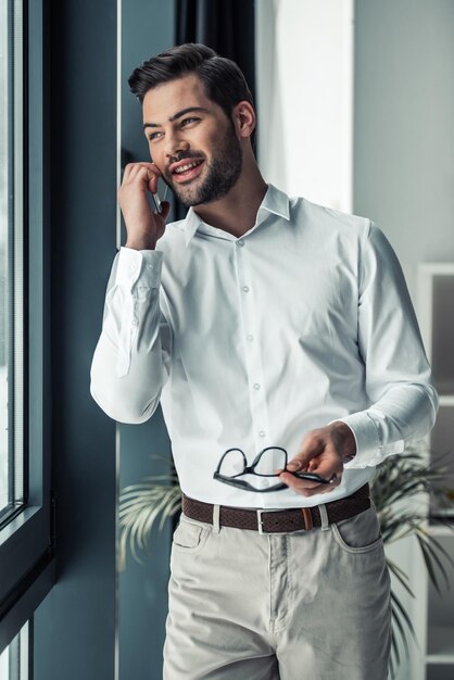 Apuesto hombre de negocios está hablando por teléfono móvil mirando por la ventana y sonriendo mientras está de pie en la oficina