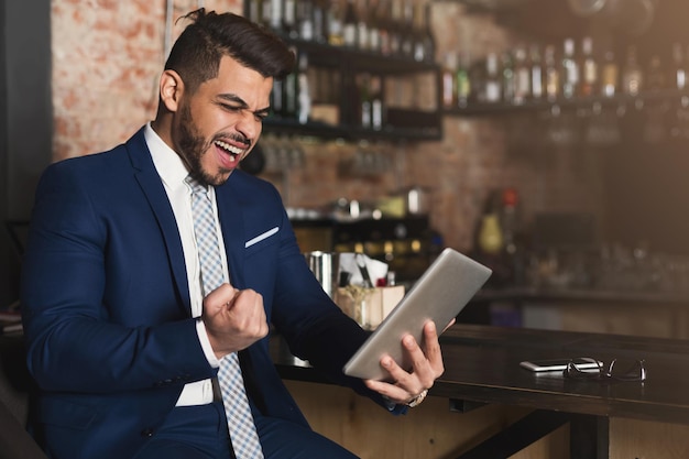 Apuesto hombre de negocios eufórico ganando y mirando a la tableta digital sentado en el mostrador de bar, espacio de copia