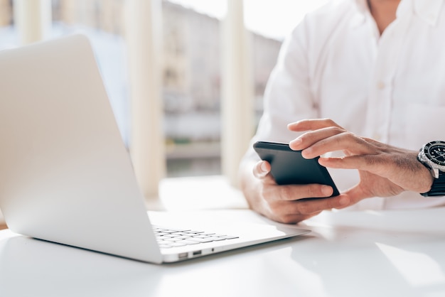 Apuesto hombre de negocios joven en una camisa blanca sentada en un café y trabajando con una computadora portátil