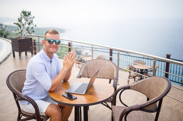 Apuesto hombre de negocios joven exitoso sentado en una mesa junto a la piscina con un portátil con vistas al mar Mediterráneo. Trabajo a distancia en vacaciones. Concepto de vacaciones