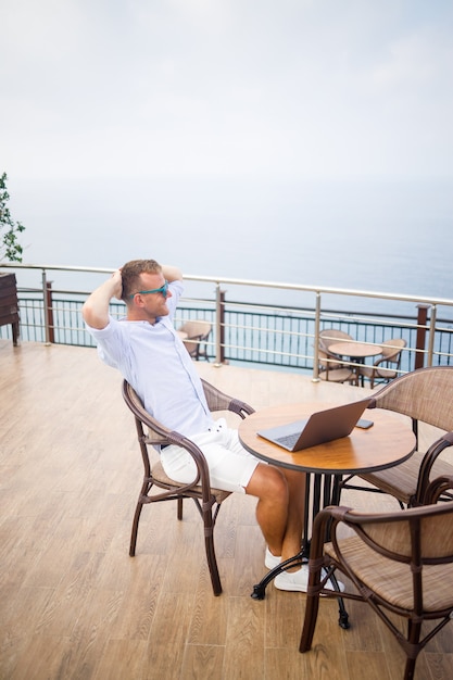 Apuesto hombre de negocios joven exitoso sentado en una mesa junto a la piscina con un portátil con vistas al mar Mediterráneo. Trabajo a distancia en vacaciones. Concepto de vacaciones