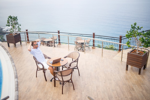 Apuesto hombre de negocios joven exitoso sentado en una mesa junto a la piscina con un portátil con vistas al mar Mediterráneo. Trabajo a distancia en vacaciones. Concepto de vacaciones