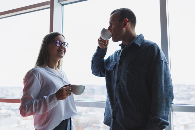 Un apuesto hombre de negocios y una joven mujer de negocios están parados y hablando en el edificio de oficinas por la mañana antes del trabajo, están bebiendo café fuerte