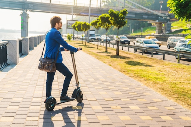 Apuesto hombre de negocios montando scooter eléctrico cerca de la carretera en la ciudad mientras hay tráfico Concepto de ecología