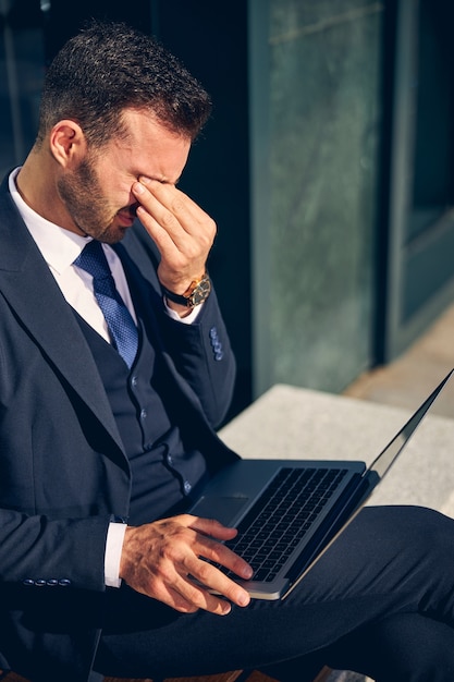 Apuesto hombre de negocios morena en ropa formal que se siente mal mientras pasa tiempo afuera solo con la computadora portátil de rodillas