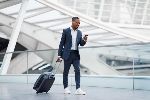 Apuesto hombre de negocios negro en el aeropuerto caminando con maleta y usando un teléfono inteligente